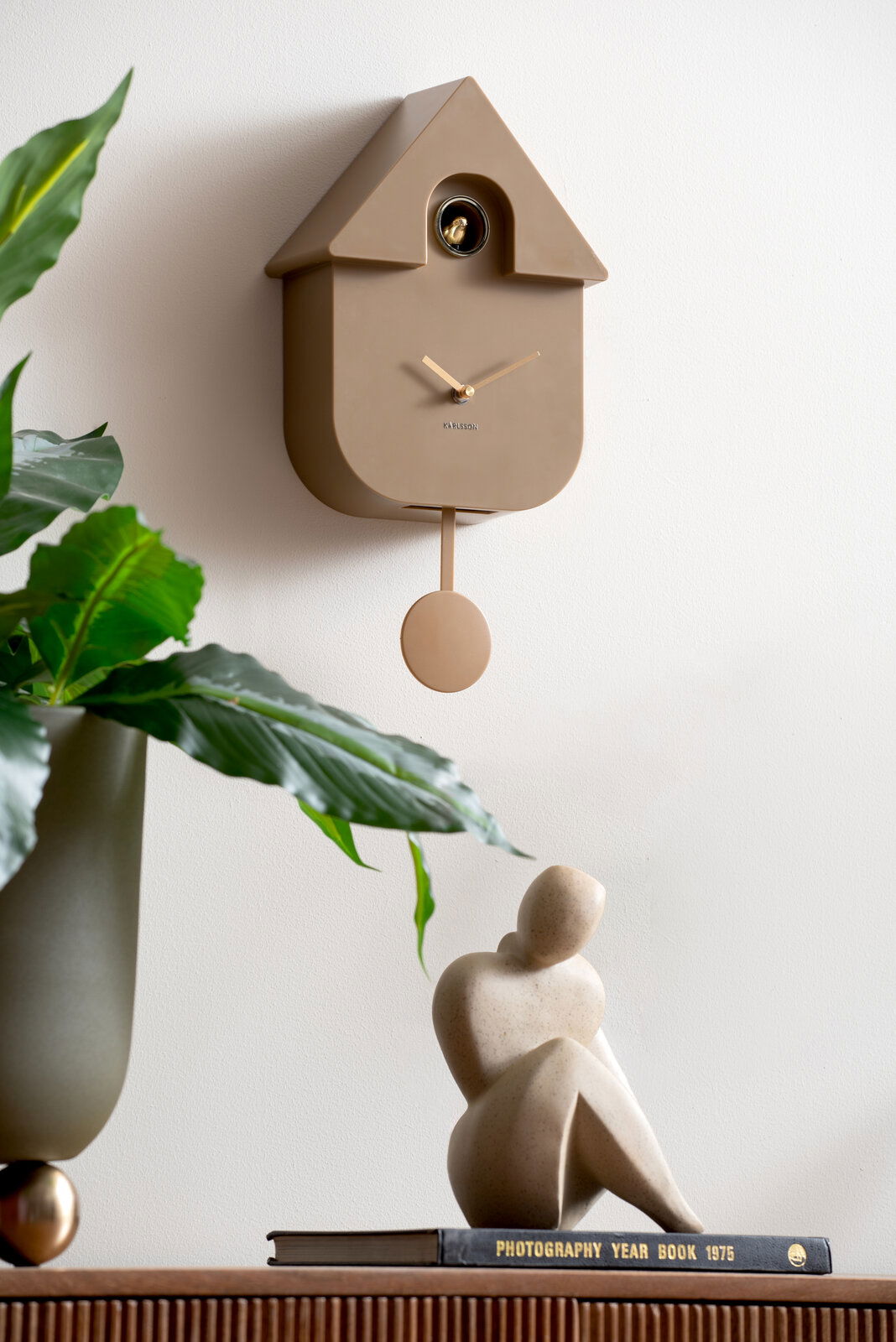 Moderne Wanduhr in Hausform mit Pendel, seitliche Perspektive, auf weißer Wand montiert, umgeben von Pflanzen und Dekoration.