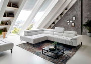 Moderne Eckgarnitur in hellem Stoffbezug, präsentiert in einem stilvollen Wohnzimmer mit schrägen Fenstern und urbanem Ausblick, aus seitlicher Perspektive fotografiert.