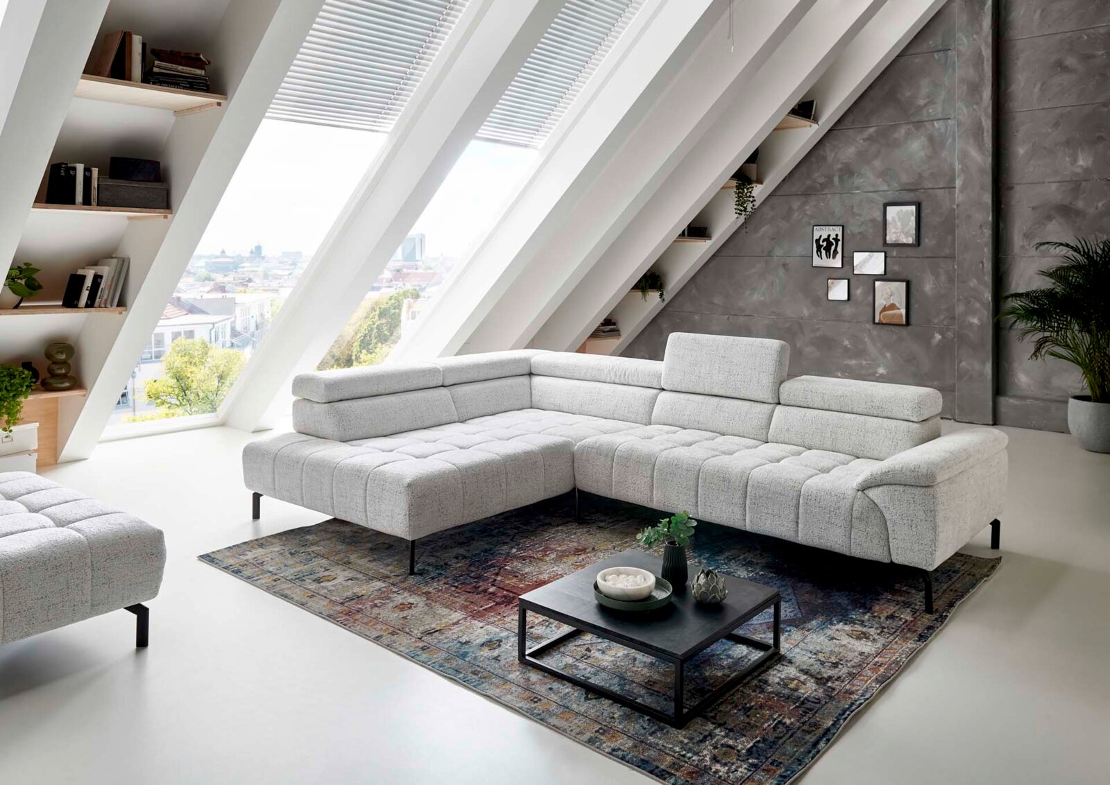Moderne Eckgarnitur in hellem Stoffbezug, präsentiert in einem stilvollen Wohnzimmer mit schrägen Fenstern und urbanem Ausblick, aus seitlicher Perspektive fotografiert.