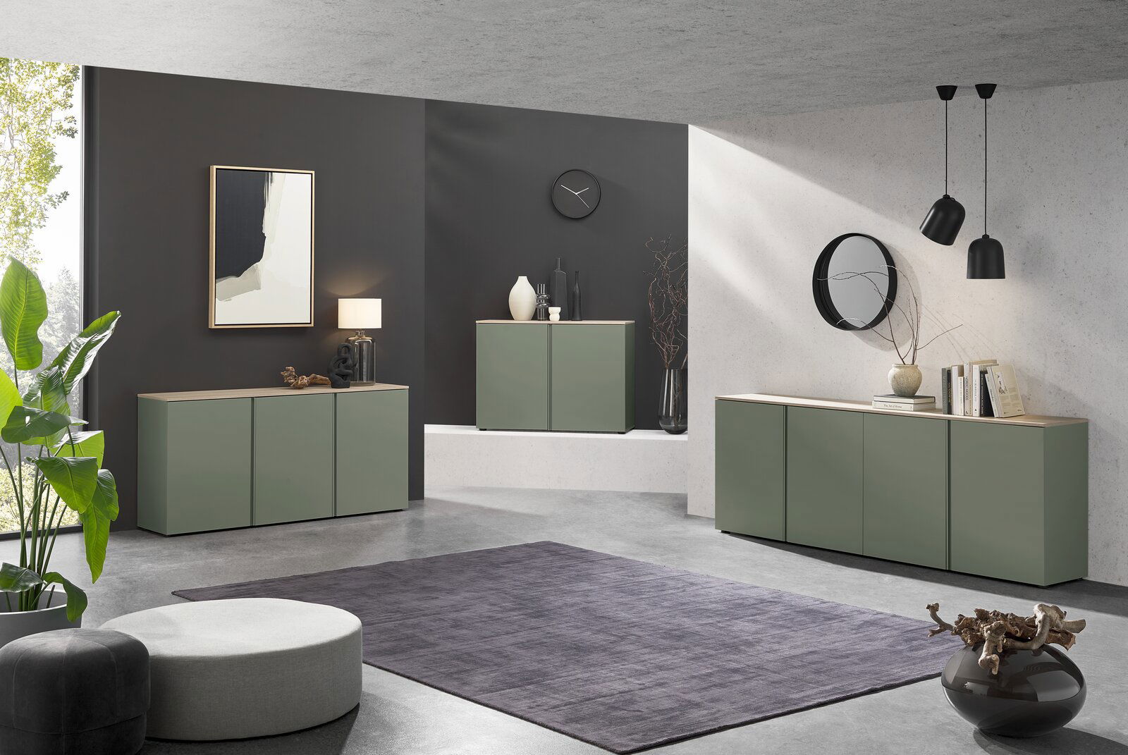 Modernes Wohnzimmer mit drei olivgrünen Sideboards mit Holzoberplatte vor dunkler und heller Wand, dekoriert mit Lampe, Vasen, Büchern und Spiegel; Perspektive: frontale Weitwinkelansicht leicht von links.