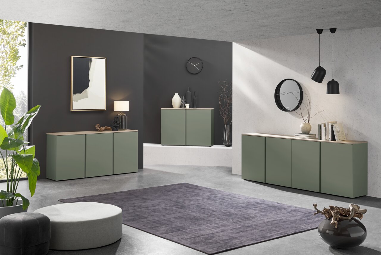 Voleo XL-Sideboard MILTON Modernes Wohnzimmer mit drei olivgrünen Sideboards mit Holzoberplatte vor dunkler und heller Wand, dekoriert mit Lampe, Vasen, Büchern und Spiegel; Perspektive: frontale Weitwinkelansicht leicht von links.