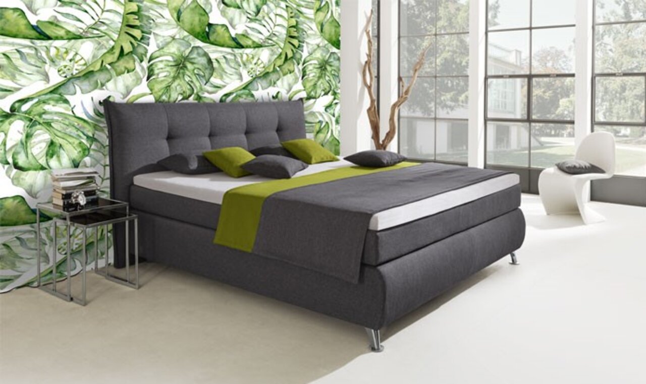 Boxspringbett in modernem Design mit grauem Stoffbezug, grünen Akzenten und mehreren Kissen, seitliche Perspektive