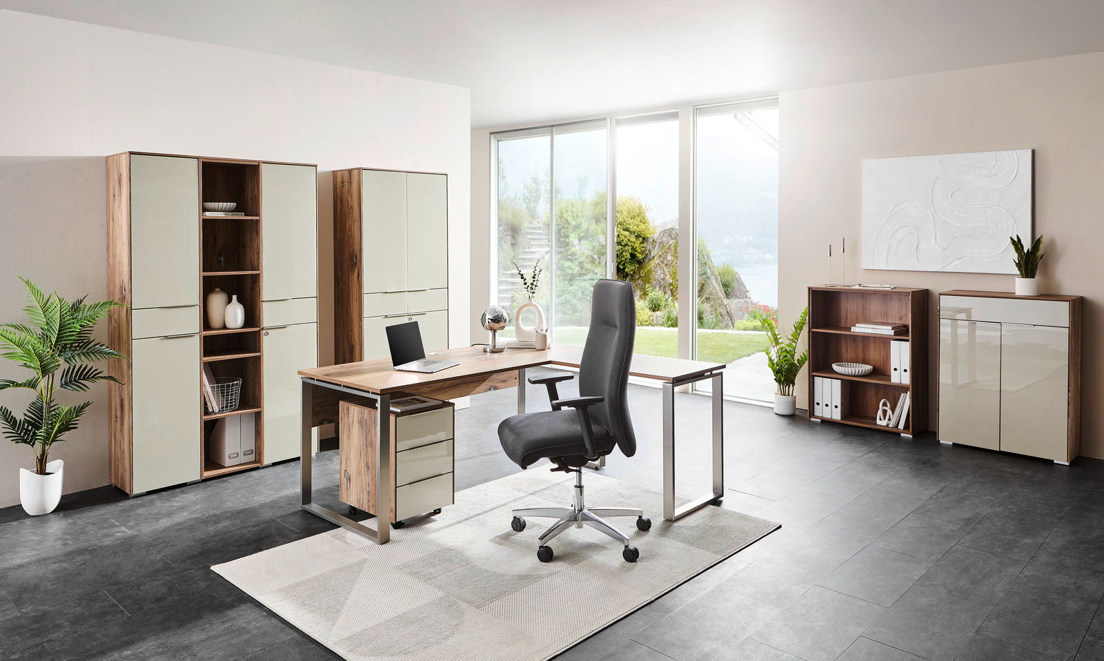 Moderne Büromöbelkombination mit Schreibtisch, Rollcontainer, Aktenschränken und Regalen in einem hellen Raum mit Blick auf den Garten, seitliche Perspektive.