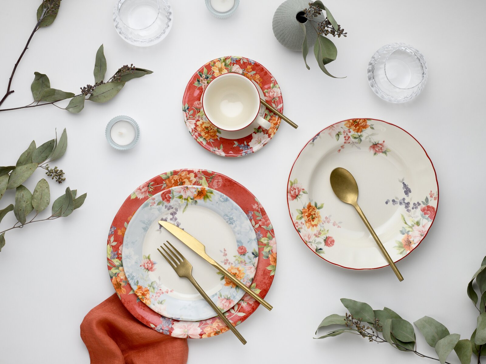 Draufsicht (Flatlay) auf ein florales Geschirr-Set: Teller und Schale mit Blumenmuster in Rot/Weiß, Tasse auf Untertasse sowie goldfarbenes Besteck auf weißem Hintergrund mit Eukalyptuszweigen und Kerzen.