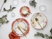 Draufsicht (Flatlay) auf ein florales Geschirr-Set: Teller und Schale mit Blumenmuster in Rot/Weiß, Tasse auf Untertasse sowie goldfarbenes Besteck auf weißem Hintergrund mit Eukalyptuszweigen und Kerzen.