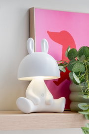LED Tischlampe in Form eines sitzenden Hasen mit Ohren, seitliche Perspektive, auf einem Regal neben einer Pflanze und einem pinken Bild.