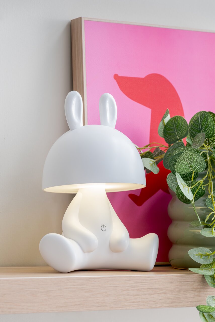 LED-tafellamp in de vorm van een zittend konijn met oren, gezien vanuit de zijkant, op een plank naast een plant en een roze schilderij.