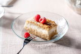 Nahaufnahme eines runden Glastellers mit einem Stück Kuchen, garniert mit Himbeeren, auf einem hellen Tisch.
