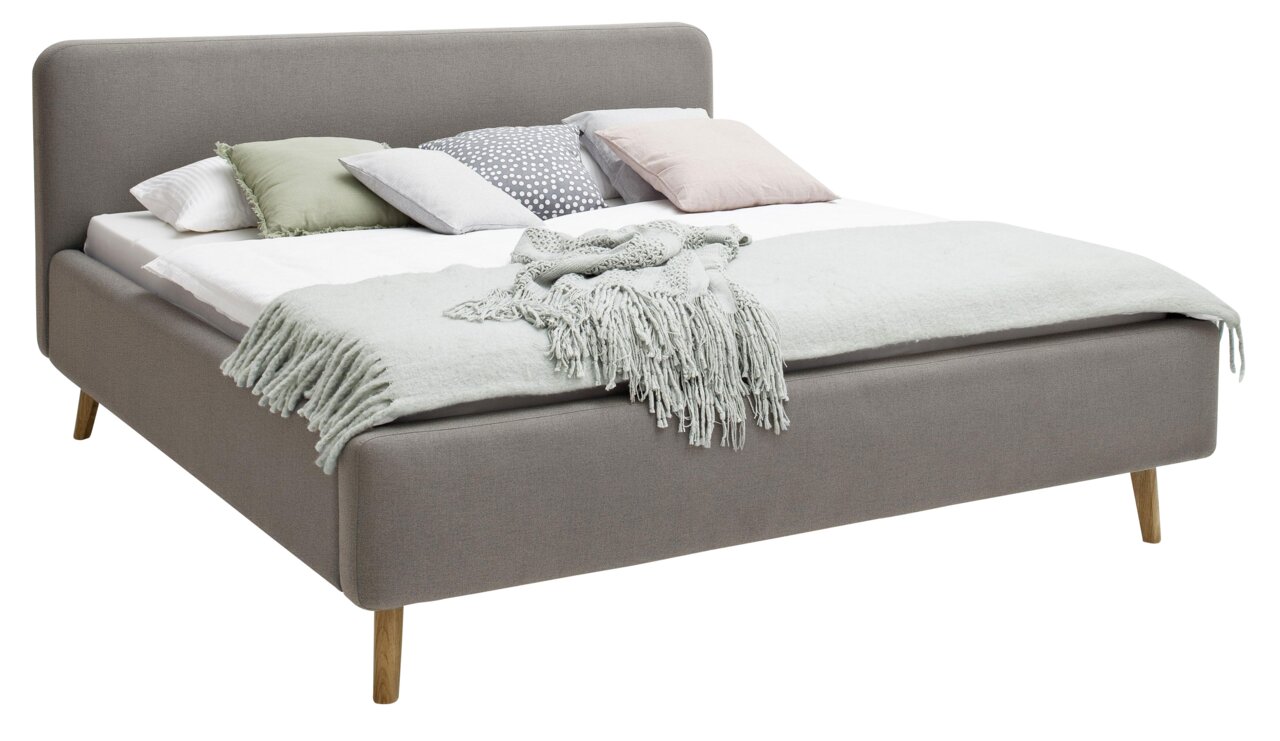Grijs gestoffeerd bed met houten poten, versierd met verschillende kussens en een deken, zijaanzicht