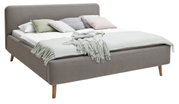 Grijs gestoffeerd bed met houten poten, versierd met verschillende kussens en een deken, zijaanzicht