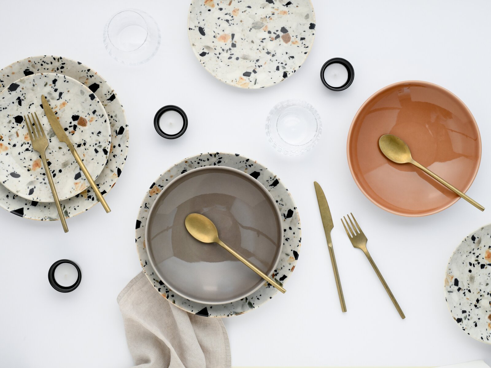 Modernes Tischgedeck mit Terrazzo-Tellern, grauer und terrakottafarbener Schale, goldfarbenem Besteck, Gläsern und Teelichtern auf weißem Hintergrund, Draufsicht von oben.