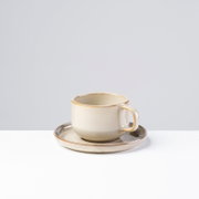 Beige Keramik-Kaffeetasse mit goldbraunem Rand auf passender Untertasse, seitliche Ansicht leicht von oben vor hellem Hintergrund