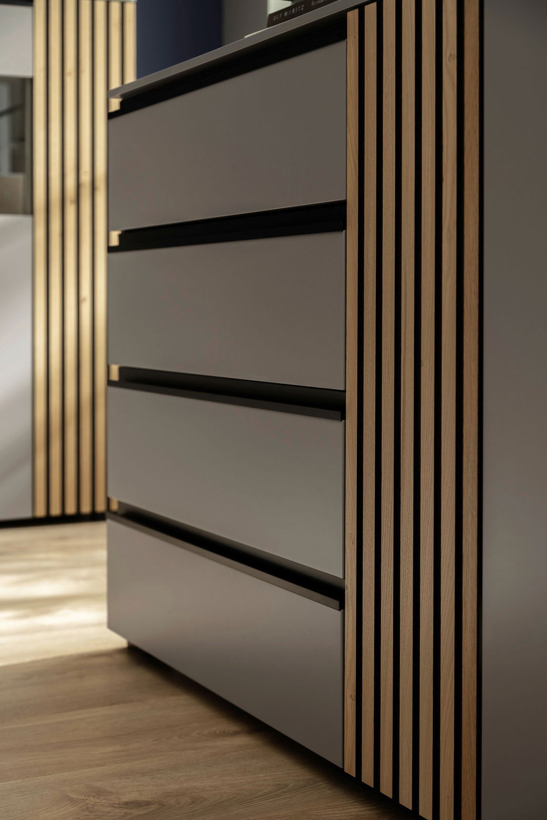 Nahaufnahme eines modernen Sideboards mit glatten, grauen Fronten und vertikalen Holzstreifen an der Seite, aus seitlicher Perspektive fotografiert.