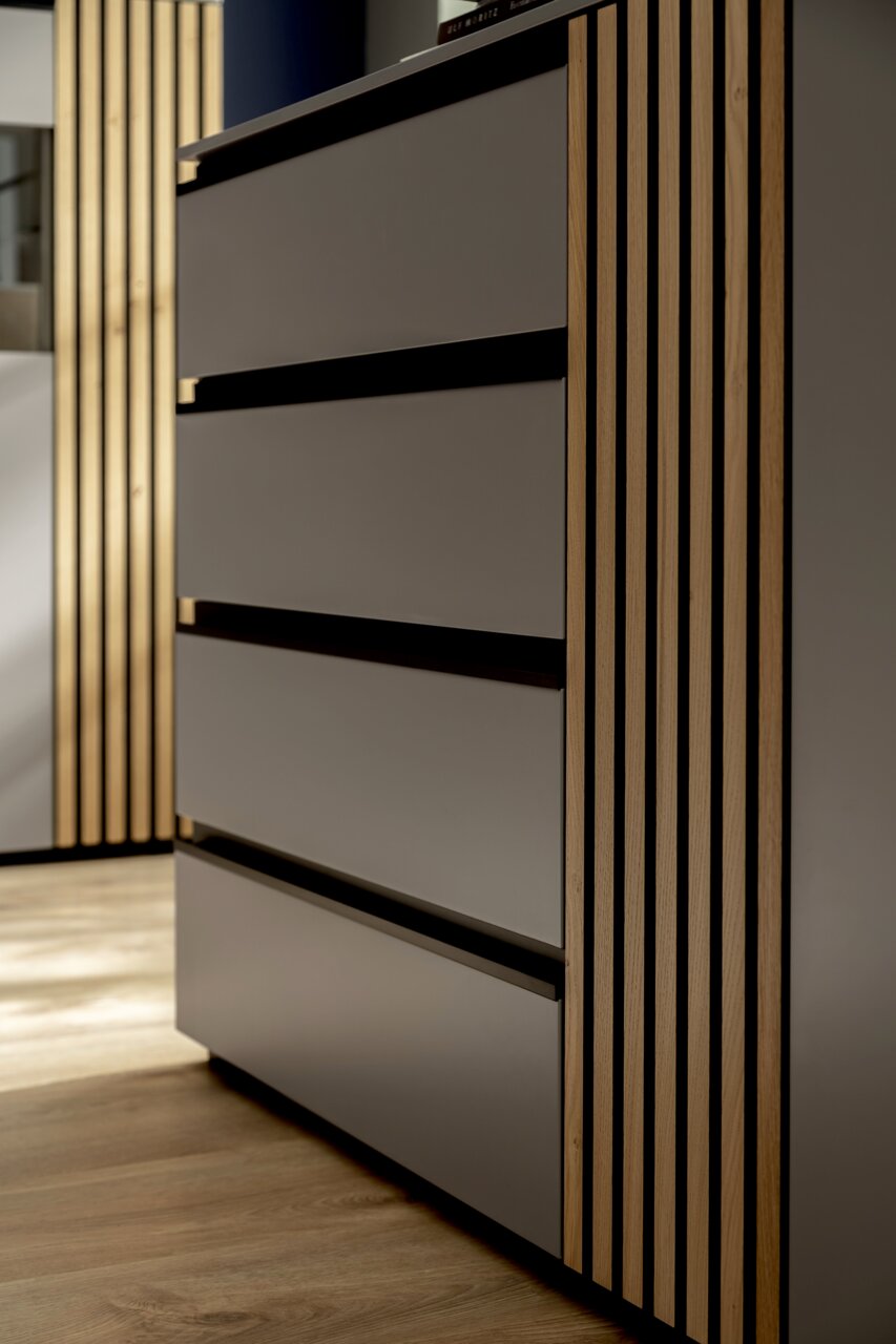 Nahaufnahme eines modernen Sideboards mit glatten, grauen Fronten und vertikalen Holzstreifen an der Seite, aus seitlicher Perspektive fotografiert.