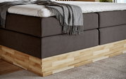 Close-up van een boxspringbed met grijze stoffen bekleding en houten onderstel, gezien vanaf de zijkant