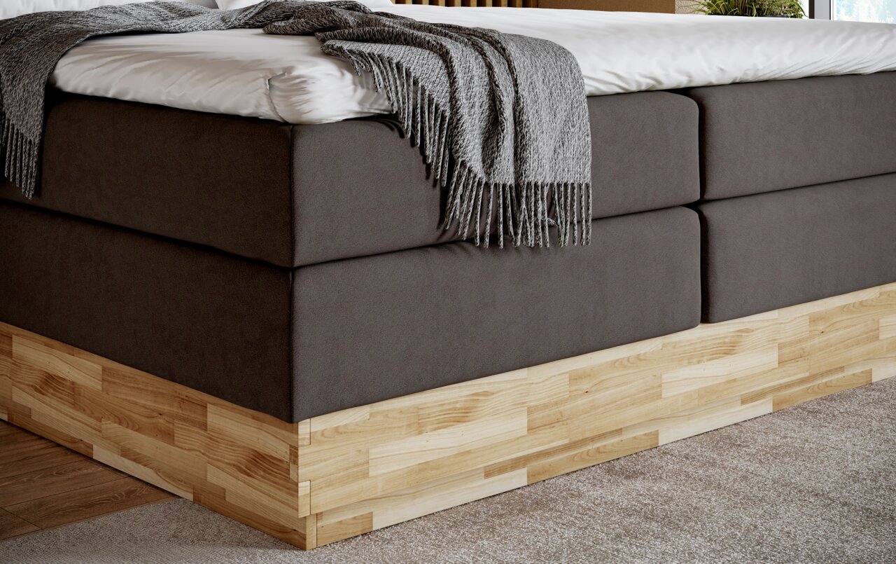 Close-up van een boxspringbed met grijze stoffen bekleding en houten onderstel, gezien vanaf de zijkant