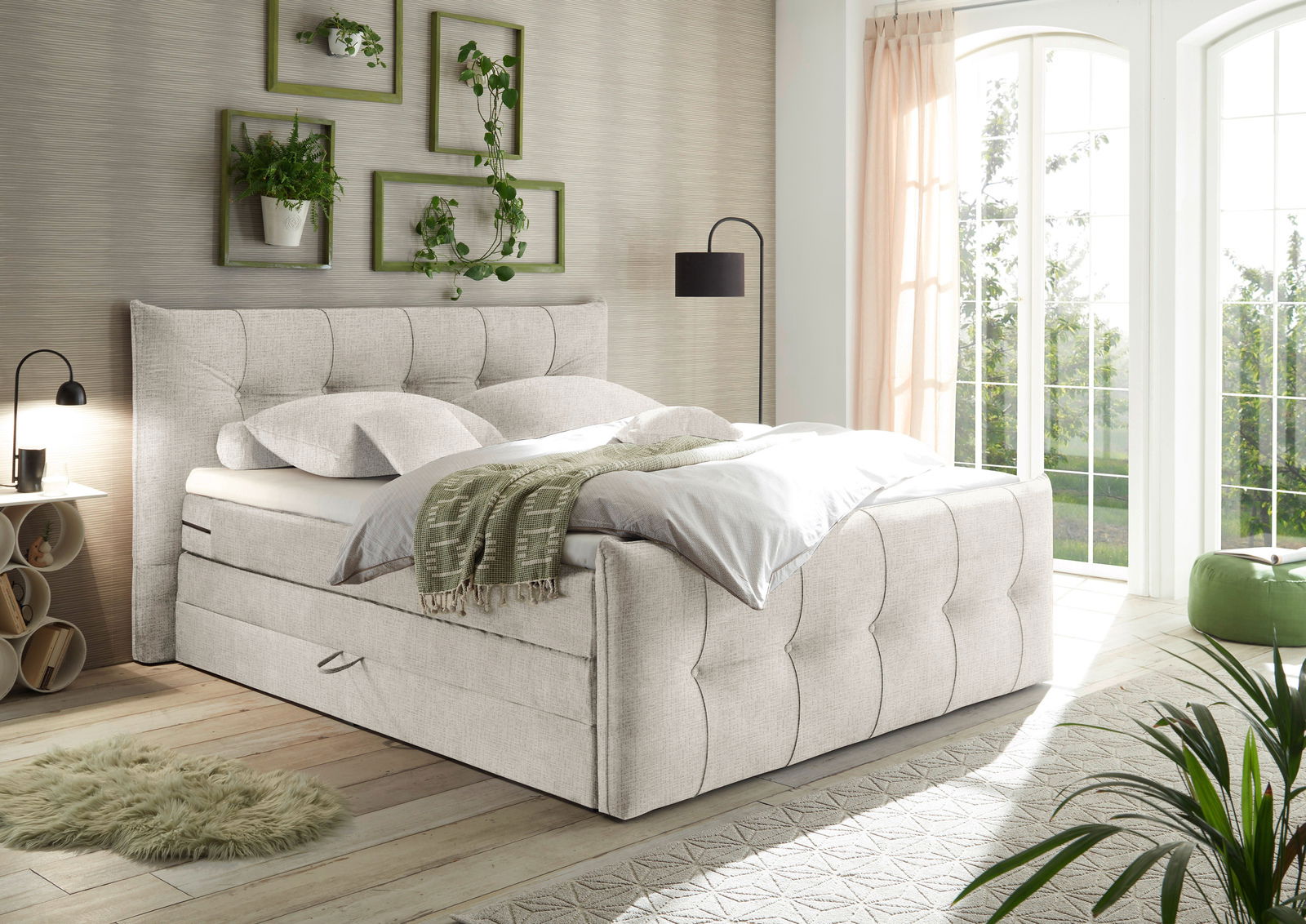 Gemütliches Boxspringbett in einem hellen Schlafzimmer mit großen Fenstern, Pflanzen und moderner Dekoration, seitliche Perspektive.