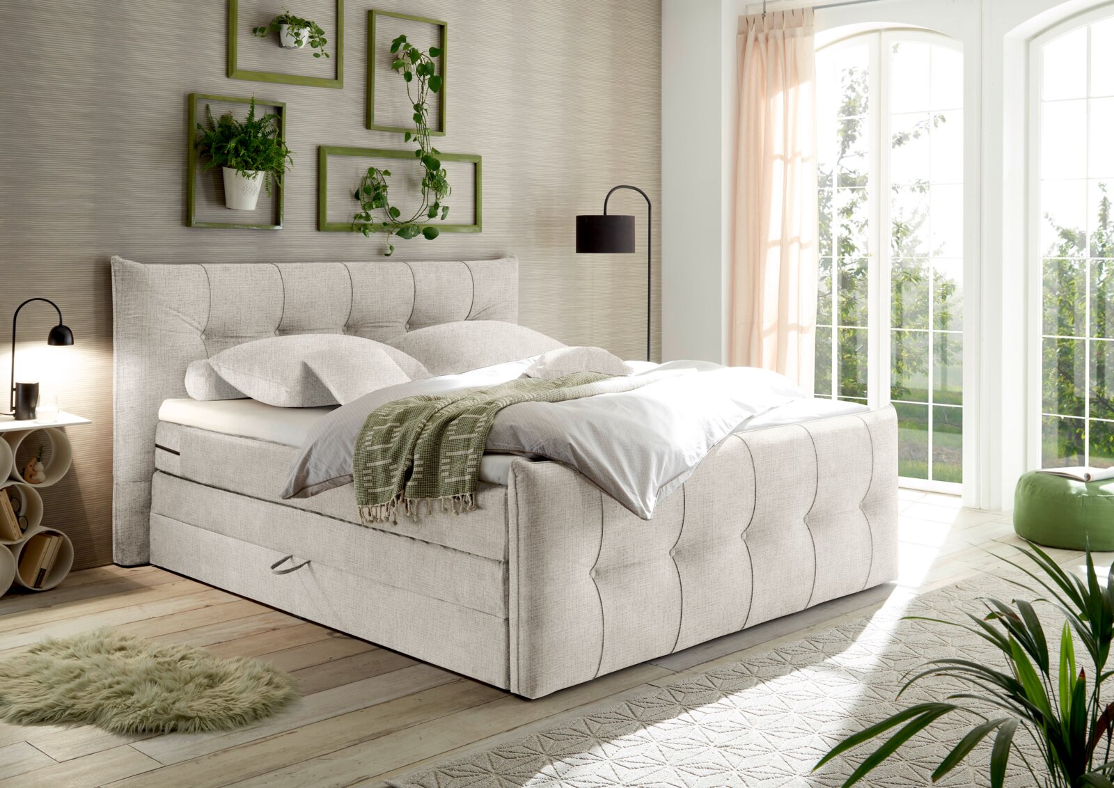 Gemütliches Boxspringbett in einem hellen Schlafzimmer mit großen Fenstern, Pflanzen und moderner Dekoration, seitliche Perspektive.