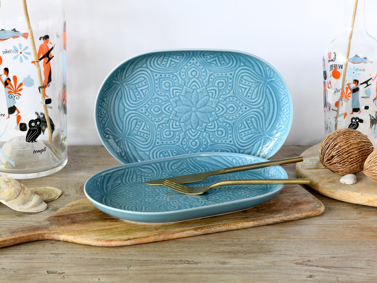 Creatable Servierteller-Set 2-lg ORIENT MANDALA Zwei ovale, türkisfarbene Keramik-Servierplatten mit geprägtem Mandala-Muster auf einem Holzbrett, vorne mit goldfarbenem Messer und Gabel; Perspektive: frontale Ansicht auf Tischhöhe.
