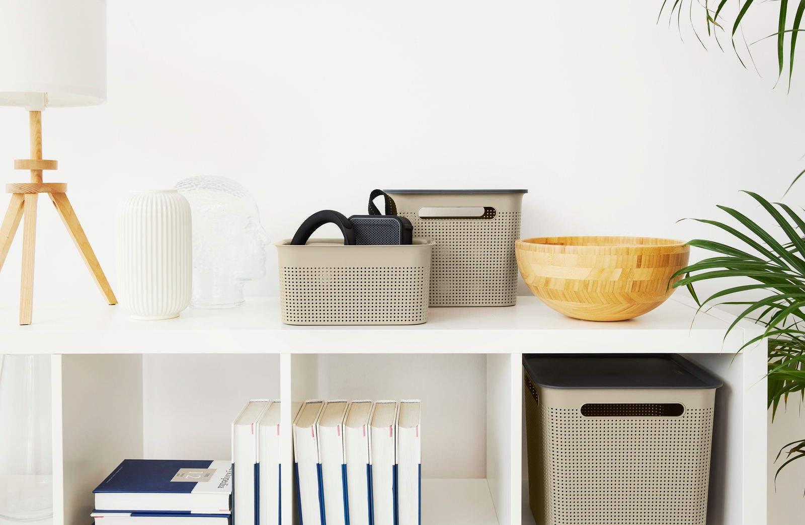 Zwei beige Aufbewahrungsboxen mit perforiertem Design, eine kleinere und eine größere, auf einem weißen Regal. Die Boxen sind von vorne sichtbar und enthalten schwarze Gegenstände. Neben den Boxen stehen dekorative Objekte wie eine Holzschale und eine wei