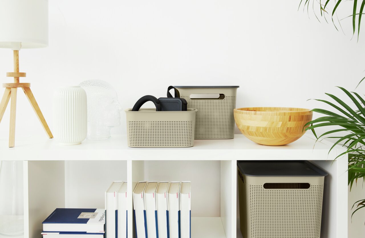 Zwei beige Aufbewahrungsboxen mit perforiertem Design, eine kleinere und eine größere, auf einem weißen Regal. Die Boxen sind von vorne sichtbar und enthalten schwarze Gegenstände. Neben den Boxen stehen dekorative Objekte wie eine Holzschale und eine wei