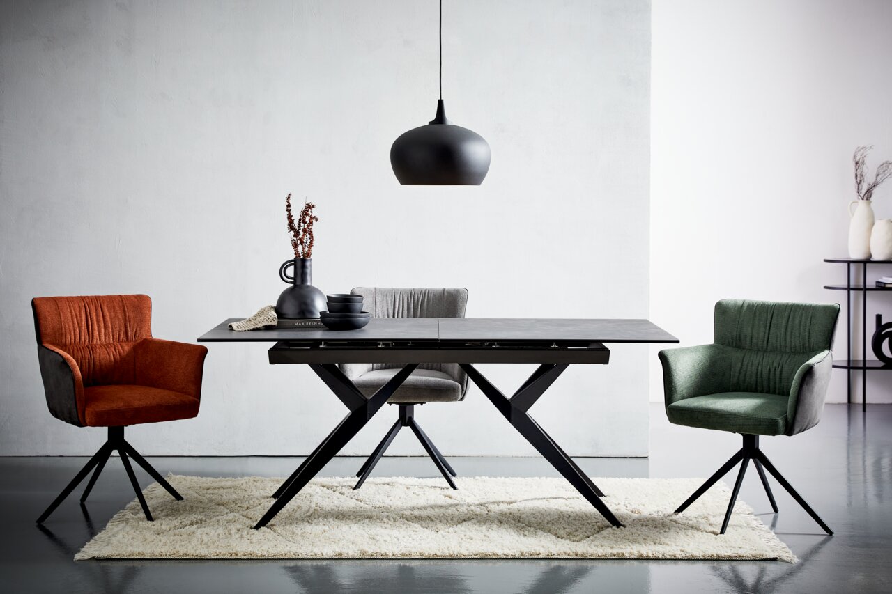Moderner Esstisch mit schwarzer Tischplatte und X-förmigen Beinen, umgeben von drei farbigen Stühlen in einem hellen Raum, aus der Frontalperspektive.