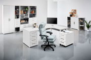 Moderne Büroumgebung mit weißen Möbeln, darunter ein Schreibtisch, Rollcontainer und Regale, aus der Perspektive eines offenen Raumes betrachtet.