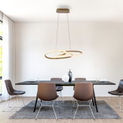 Moderne LED-Pendelleuchte mit zwei ineinander verschlungenen, ringförmigen Lichtbändern in Goldoptik über einem Esstisch, frontal aus leicht erhöhter Perspektive aufgenommen