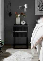 Moderne schwarze Nachtkommode mit zwei Schubladen, dekoriert mit einer Vase und einer Uhr, seitliche Perspektive.