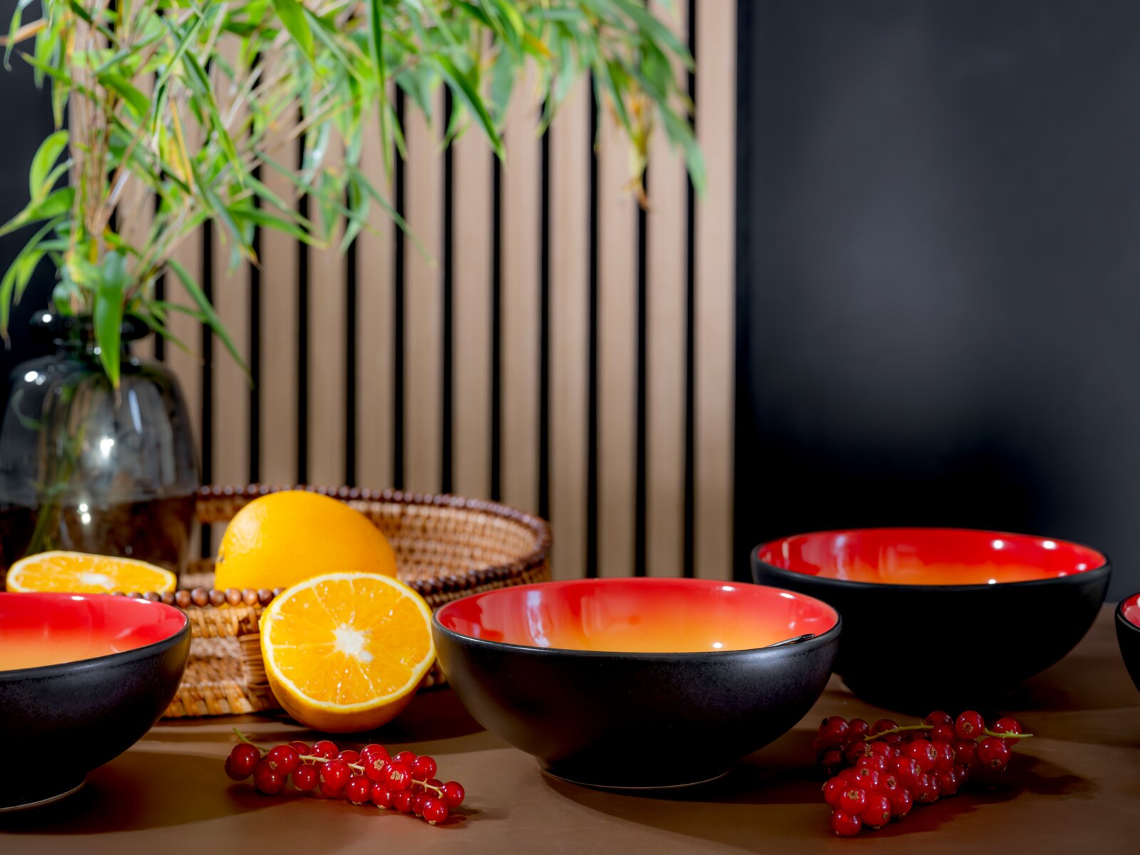 Schwarze Keramikschalen mit rot-orangefarbener Innenglasur, aus leicht erhöhter Frontperspektive zwischen Orangen, roten Johannisbeeren und dekorativem Grün arrangiert