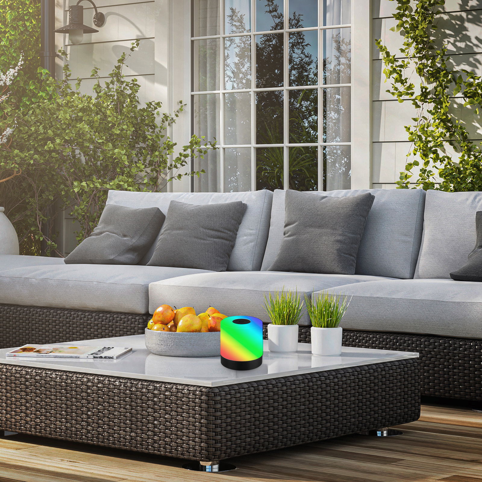 Akku RGB LED Tischleuchte auf einem modernen Terrassentisch, umgeben von einem gemütlichen Sofa und Pflanzen, aus der Perspektive von vorne.