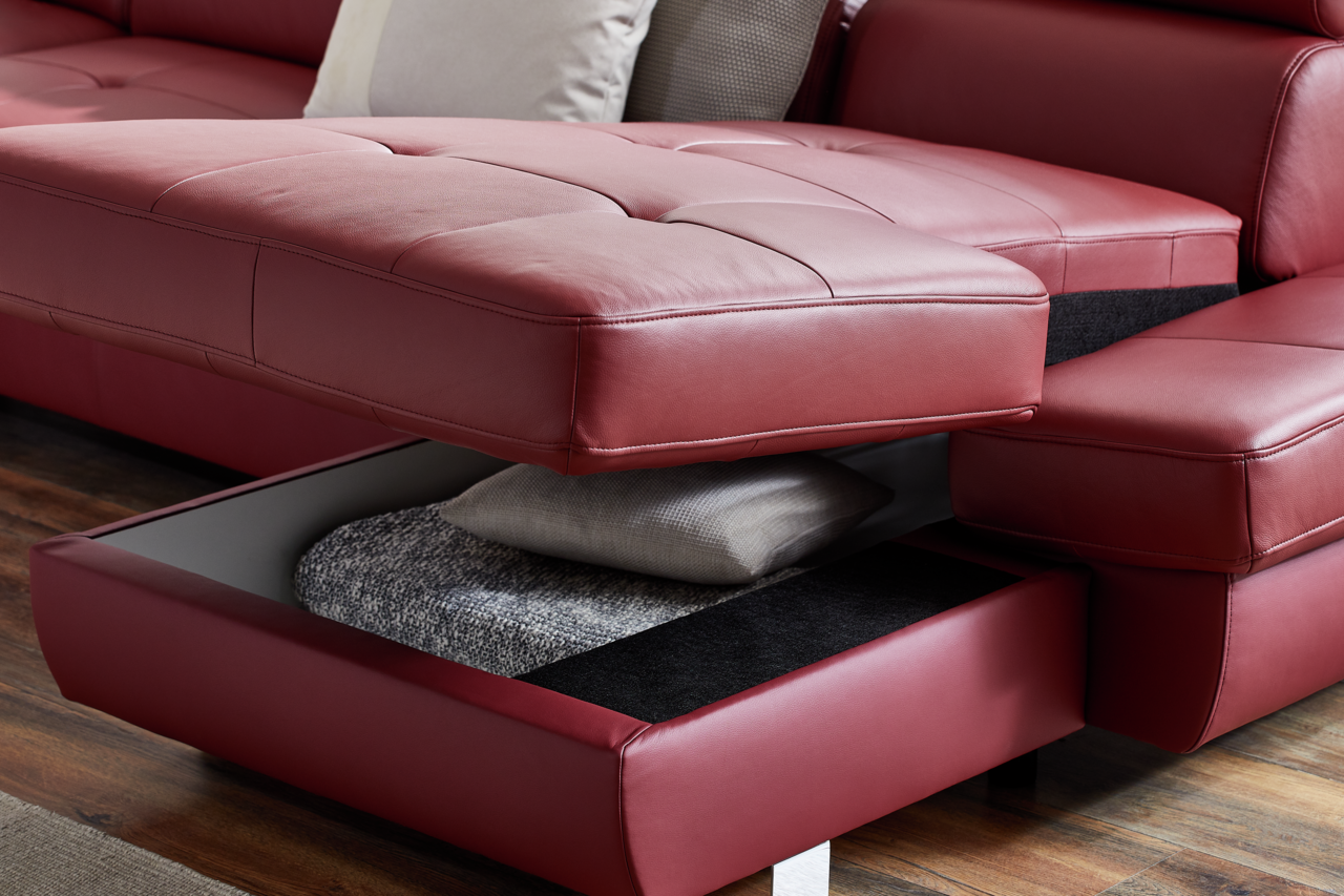 Nahaufnahme einer roten Polsterrundecke TORINO mit Bettfunktion, die einen geöffneten Stauraum mit Kissen und Decke zeigt, aus seitlicher Perspektive.