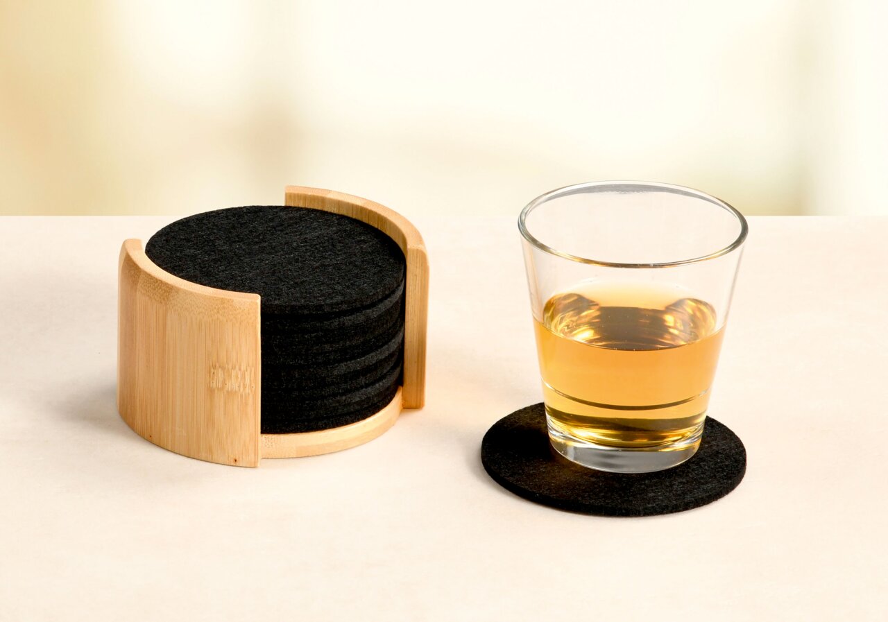 Ein Set aus schwarzen Filzuntersetzern in einem Holzhalter, mit einem Glas auf einem Untersetzer. Perspektive: seitlich.