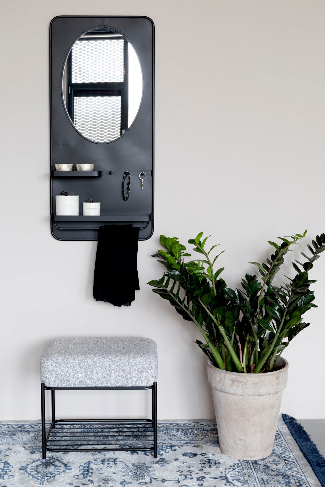 Z2 Hocker MILOU Moderner Hocker mit grauem Polster und schwarzem Metallgestell, frontal fotografiert, neben einem großen grünen Pflanzenkübel auf einem gemusterten Teppich.