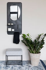 Moderner Hocker mit grauem Polster und schwarzem Metallgestell, frontal fotografiert, neben einem großen grünen Pflanzenkübel auf einem gemusterten Teppich.