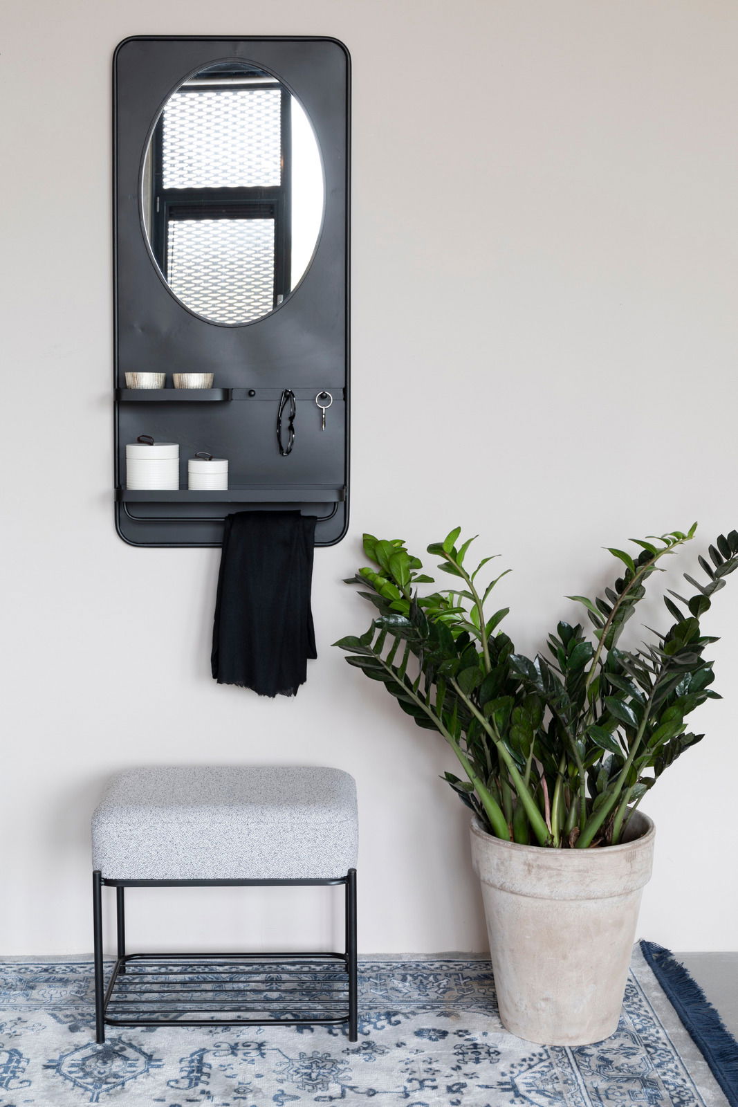 Z2 Hocker MILOU Moderner Hocker mit grauem Polster und schwarzem Metallgestell, frontal fotografiert, neben einem großen grünen Pflanzenkübel auf einem gemusterten Teppich.