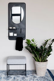 Moderner Hocker mit grauem Polster und schwarzem Metallgestell, frontal fotografiert, neben einem großen grünen Pflanzenkübel auf einem gemusterten Teppich.