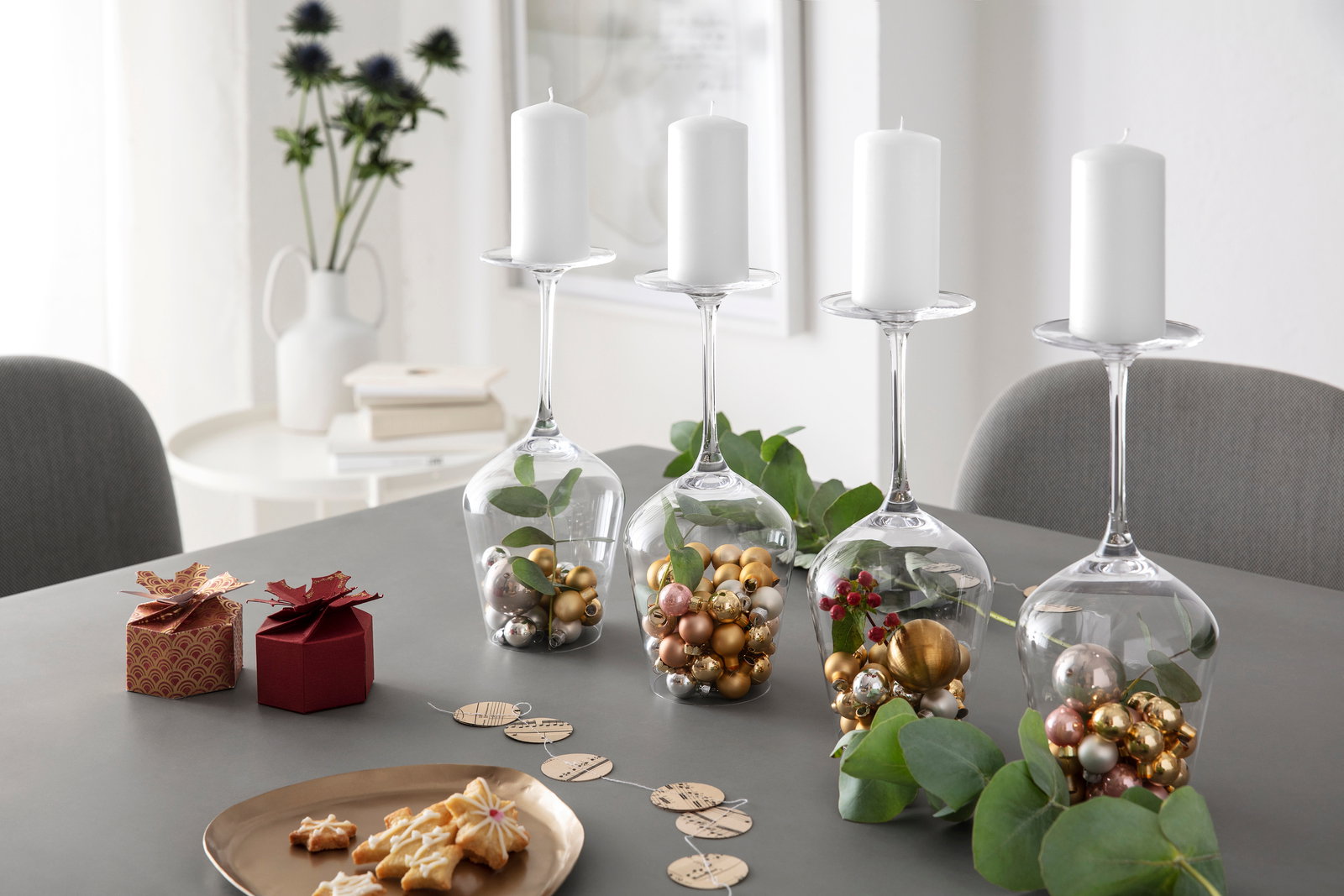 Vier umgedrehte Bordeauxkelche auf einem Tisch, gefüllt mit goldenen und silbernen Kugeln sowie grünen Blättern, dienen als Kerzenhalter für weiße Stumpenkerzen. Perspektive: seitlich von vorne.