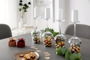 Vier omgekeerde bordeauxrode kelken op een tafel, gevuld met gouden en zilveren bolletjes en groene blaadjes, dienen als kandelaars voor witte stompkaarsen. Perspectief: zijaanzicht vanaf de voorkant.