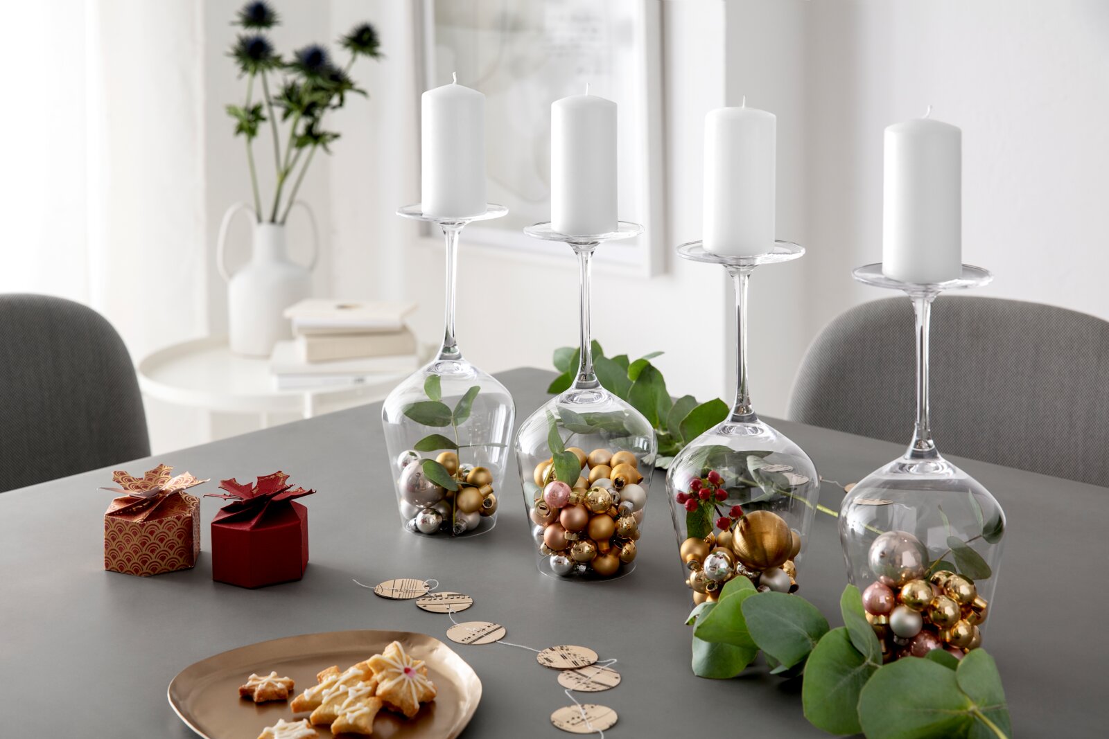 Vier omgekeerde bordeauxrode kelken op een tafel, gevuld met gouden en zilveren bolletjes en groene blaadjes, dienen als kandelaars voor witte stompkaarsen. Perspectief: zijaanzicht vanaf de voorkant.