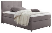 Boxspringbett in seitlicher Perspektive mit grauem Stoffbezug, Kopfteil mit quadratischer Steppung, und dekorativen Kissen und Decke.