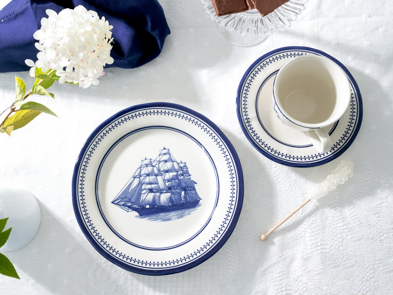 Draufsicht auf blau-weißes Porzellan-Geschirr: Teller mit Segelschiff-Motiv und Tasse auf Untertasse auf weißer Tischdecke, dekoriert mit Blume und Zuckerstange.