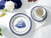 Draufsicht auf blau-weißes Porzellan-Geschirr: Teller mit Segelschiff-Motiv und Tasse auf Untertasse auf weißer Tischdecke, dekoriert mit Blume und Zuckerstange.
