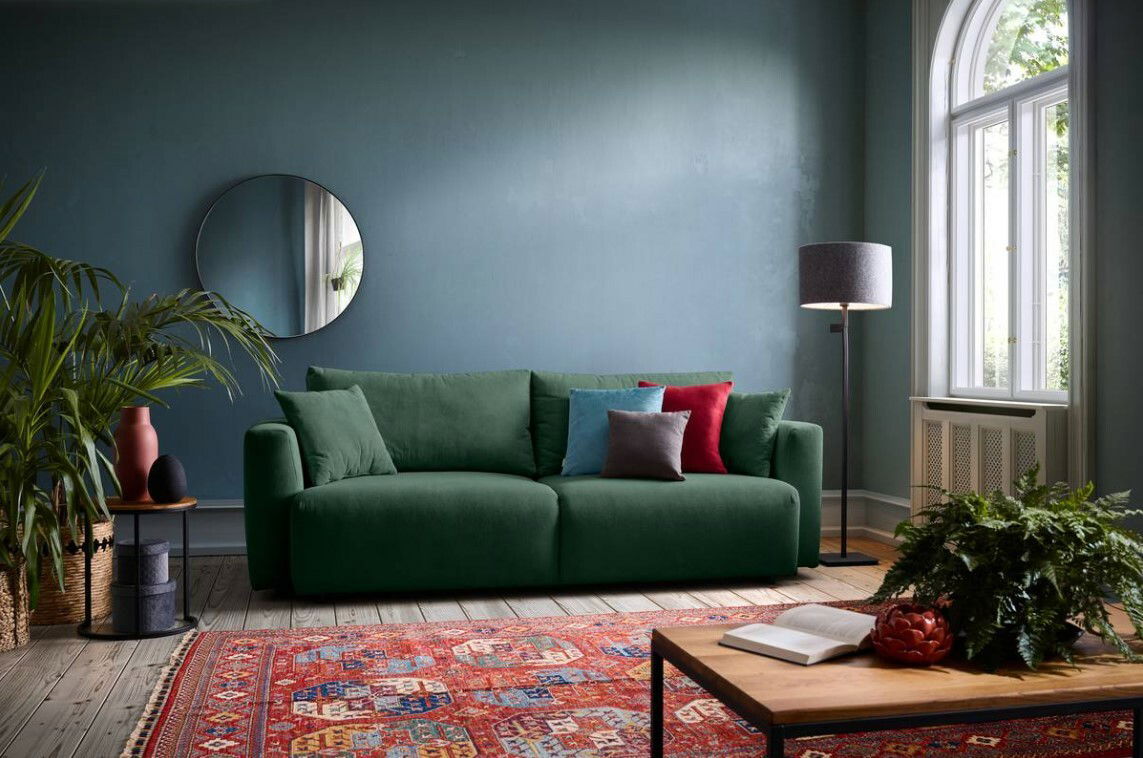 Grünes Funktionssofa in einem stilvoll eingerichteten Wohnzimmer, frontal betrachtet, mit bunten Kissen und einem runden Spiegel an der Wand.