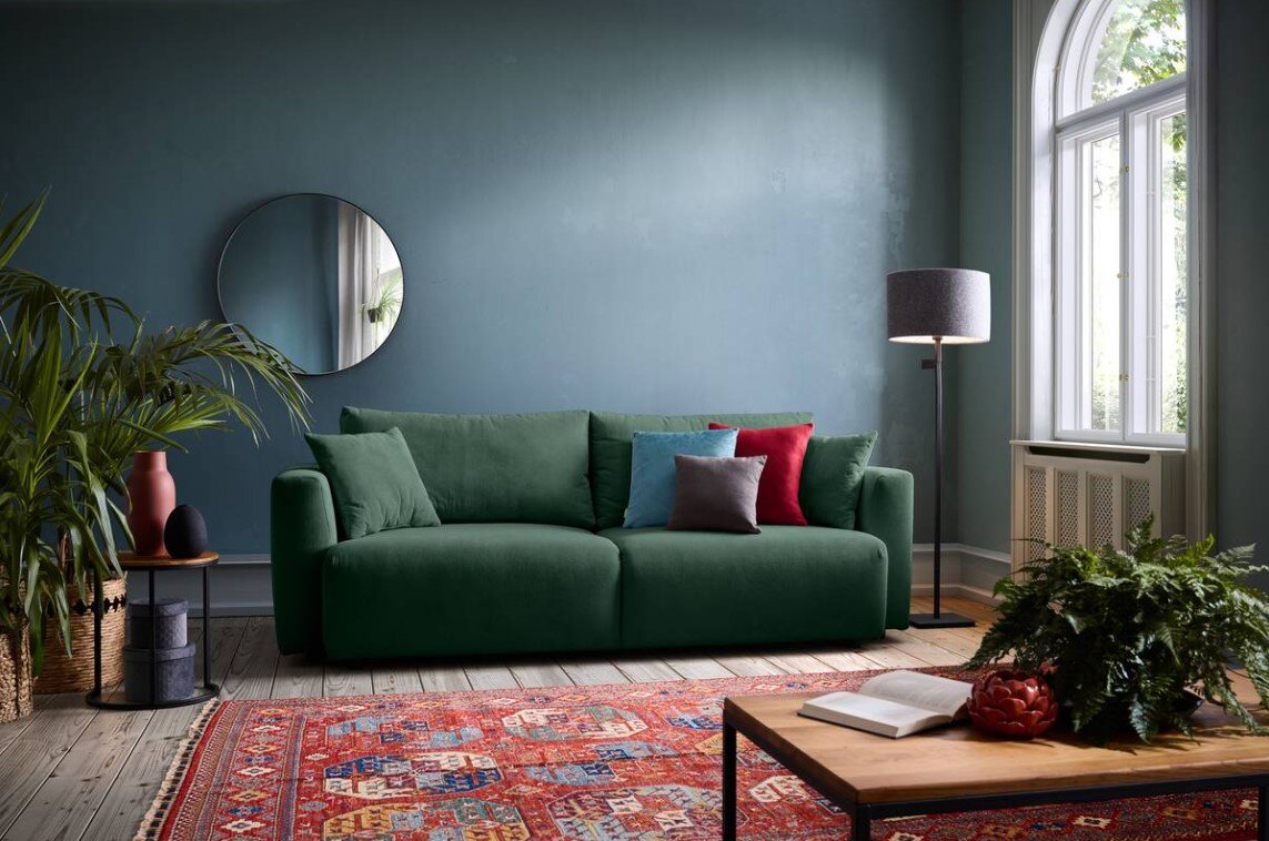 Grünes Funktionssofa in einem stilvoll eingerichteten Wohnzimmer, frontal betrachtet, mit bunten Kissen und einem runden Spiegel an der Wand.