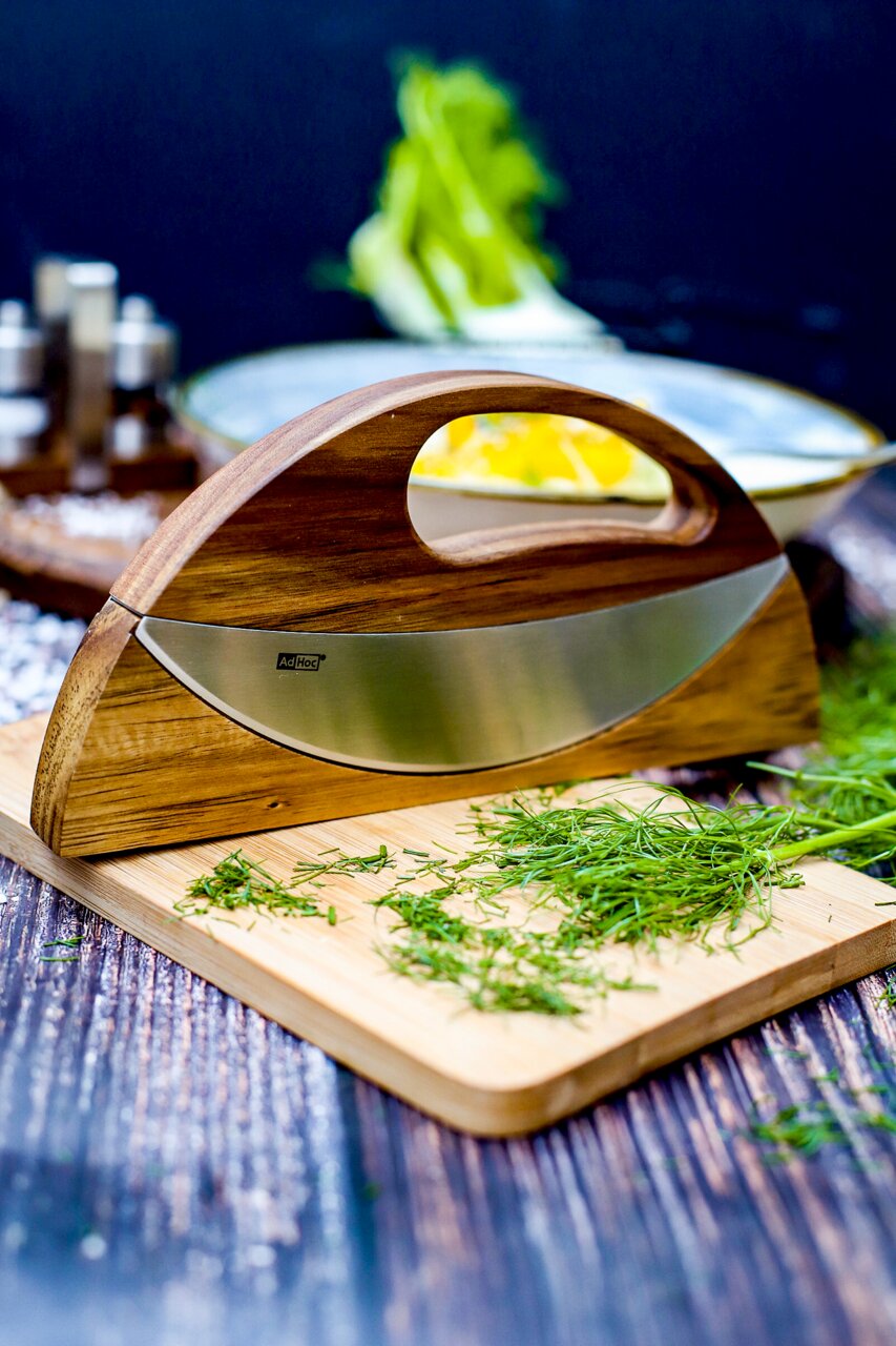 Doppel-Wiegemesser WAVE aus Holz und Edelstahl, seitliche Perspektive, auf einem Schneidebrett mit frischen Kräutern.