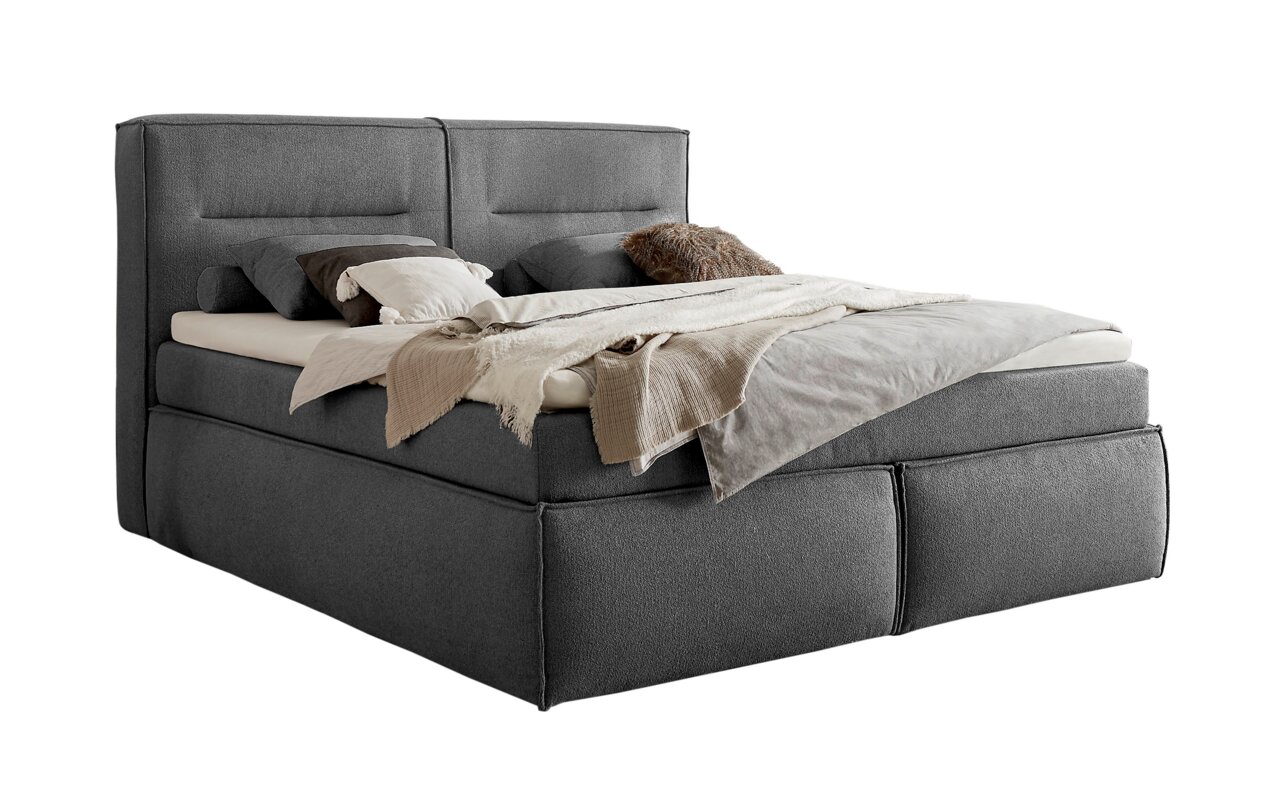 Graues Boxspringbett mit Kissen und Decken, seitliche Perspektive