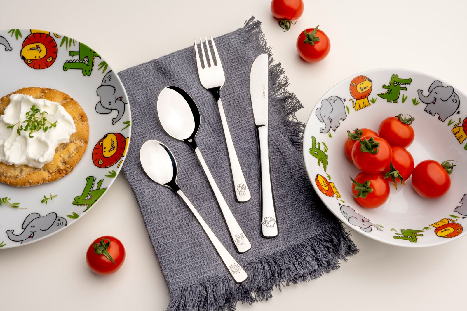 Kinderbesteck 4-tlg. mit Tiermotiven, bestehend aus Messer, Gabel, großer und kleiner Löffel, auf grauem Tuch arrangiert, umgeben von bunten Kindertellern mit Tiermotiven und frischen Tomaten, aus der Vogelperspektive fotografiert.
