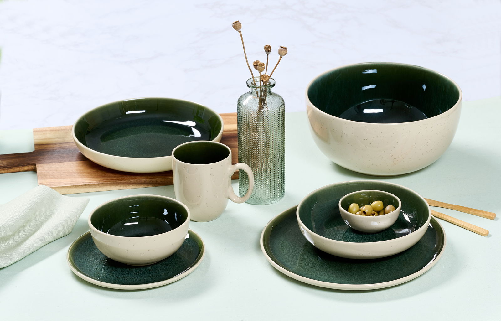 Ritzenhoff & Breker Becher SKOG Vielseitiges Geschirrset in elegantem Dunkelgrün und Creme, bestehend aus Tassen, Tellern und Schalen, auf einem Tisch arrangiert. Seitliche Perspektive.