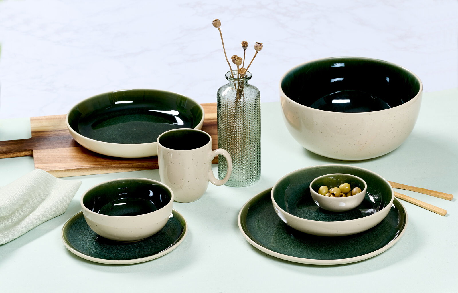 Ritzenhoff & Breker Becher SKOG Vielseitiges Geschirrset in elegantem Dunkelgrün und Creme, bestehend aus Tassen, Tellern und Schalen, auf einem Tisch arrangiert. Seitliche Perspektive.